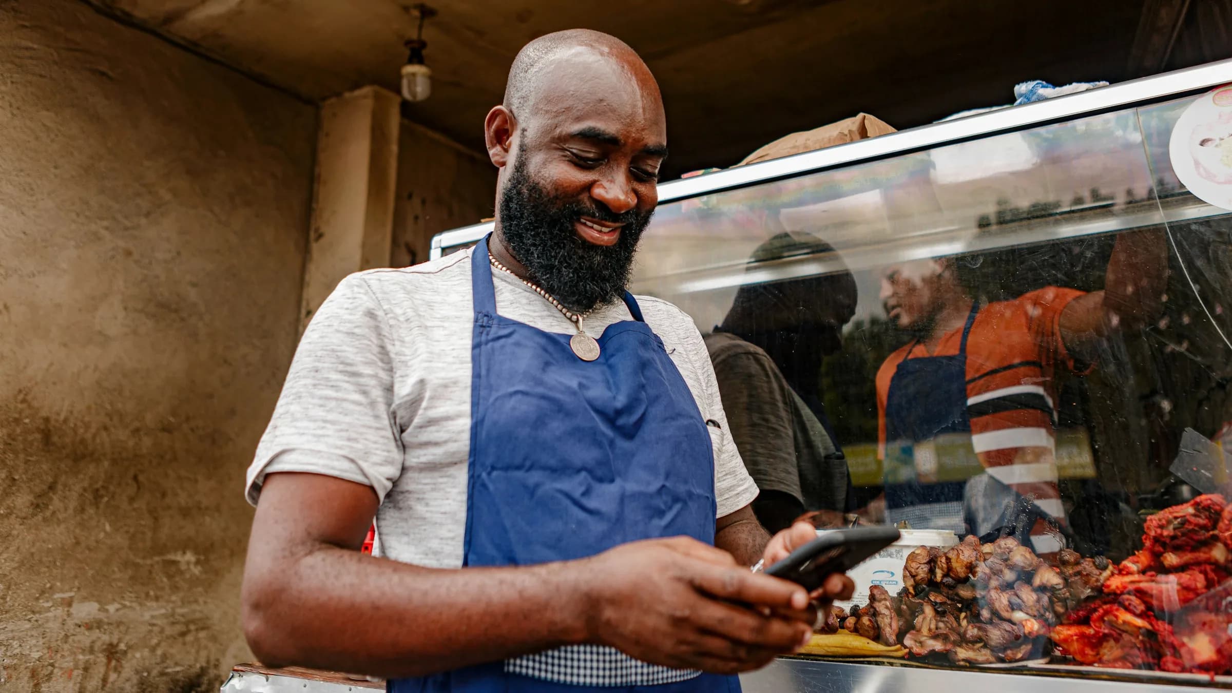 A butcher in a blue apron smiles at his phone behind a counter of cooked meats.