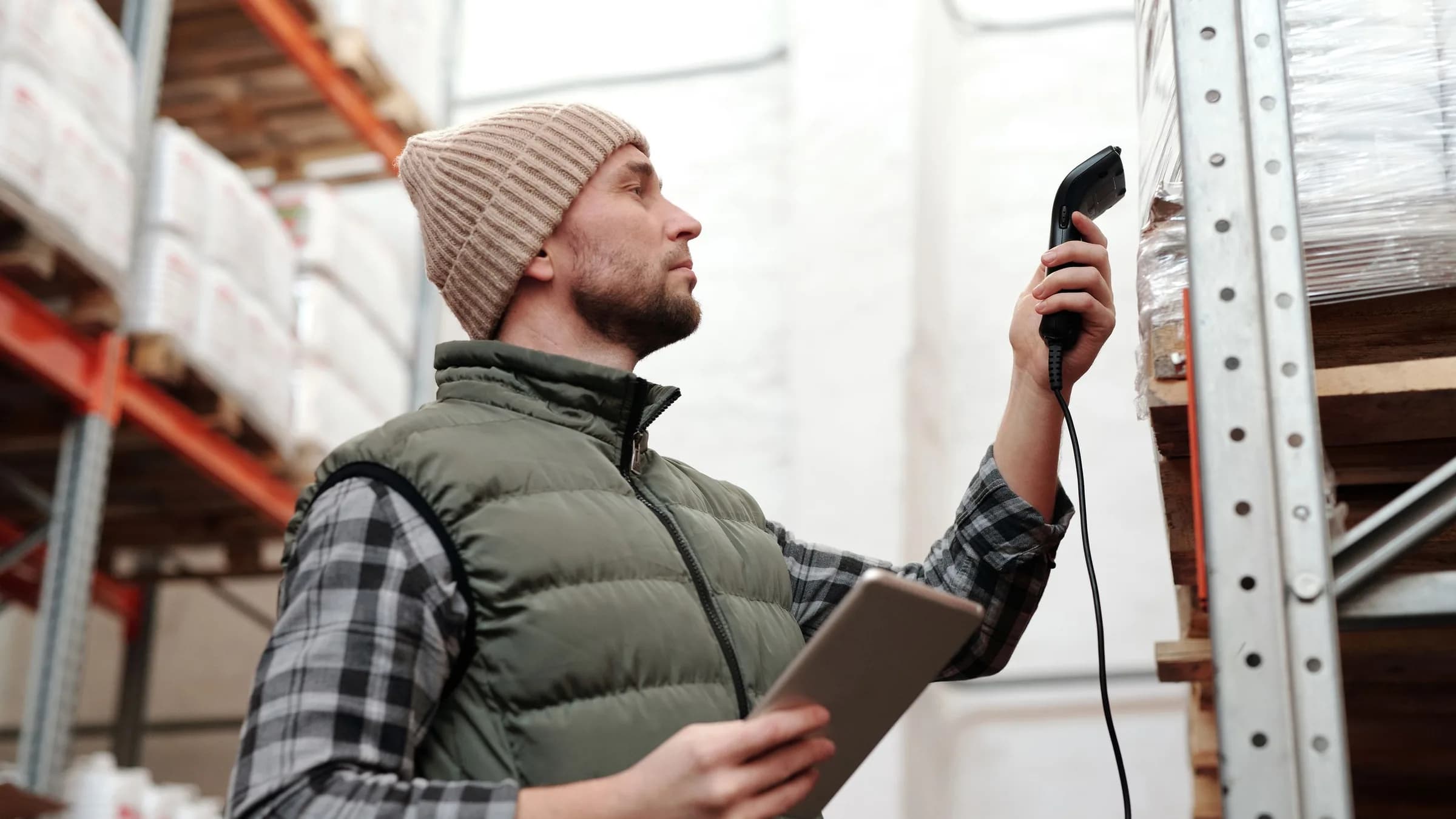A stockroom worker in a beanie scans a shelf with a corded handheld scanner, a tablet in his other hand.