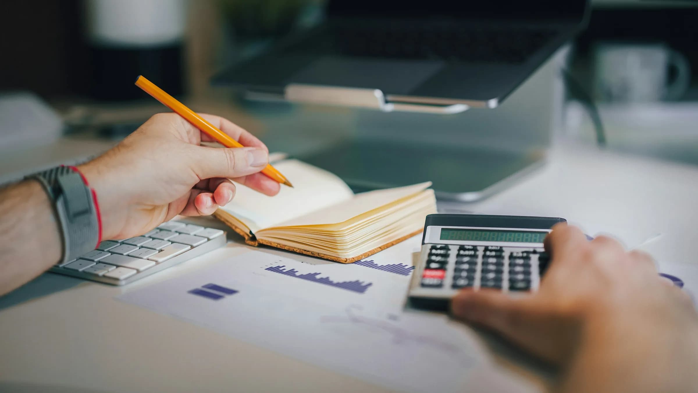 Hands working through the numbers with a pencil, a small notebook and a calculator on a desk.