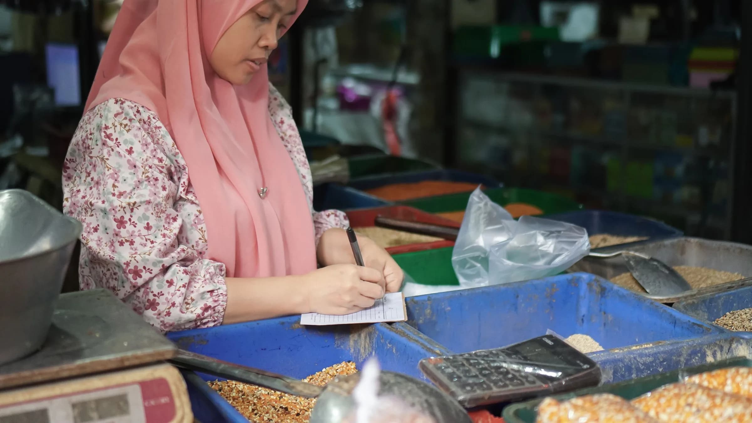 A market stallholder writes in a small notebook at her counter, a calculator beside the trays of grain.