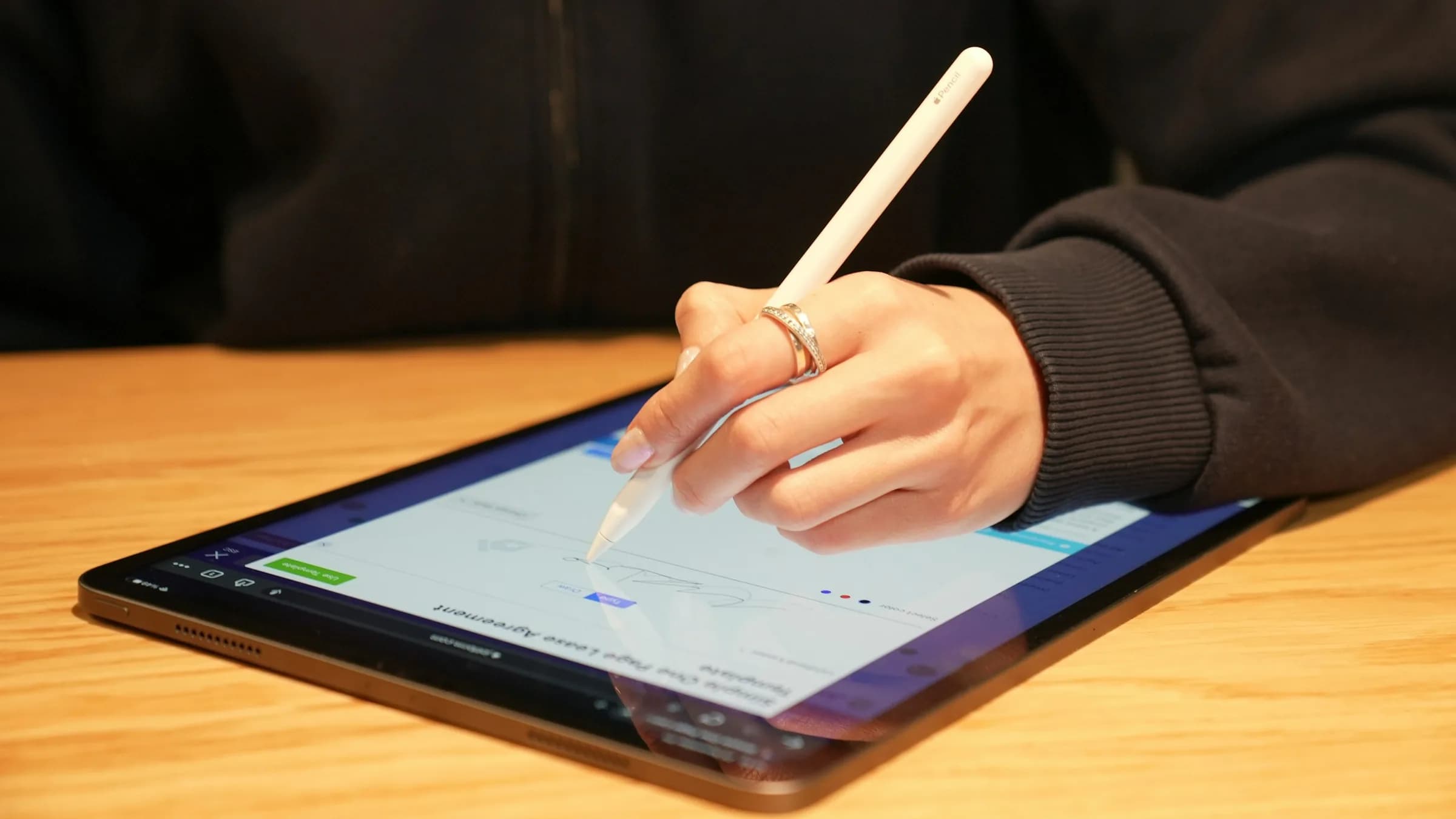 A hand signs a document on a tablet with a stylus at a wooden table.