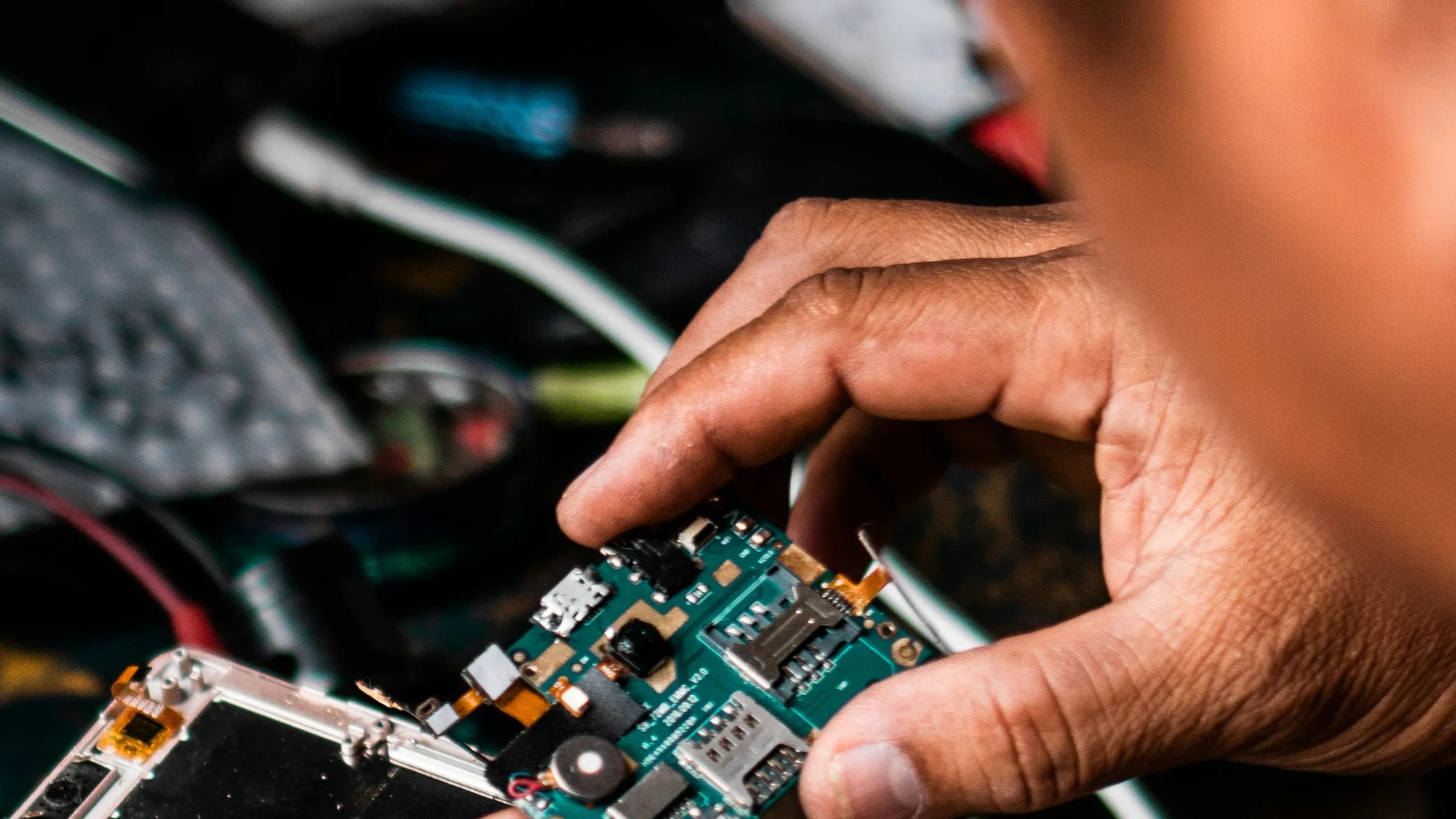 A technician holds the circuit board of an opened smartphone over a cluttered repair bench.