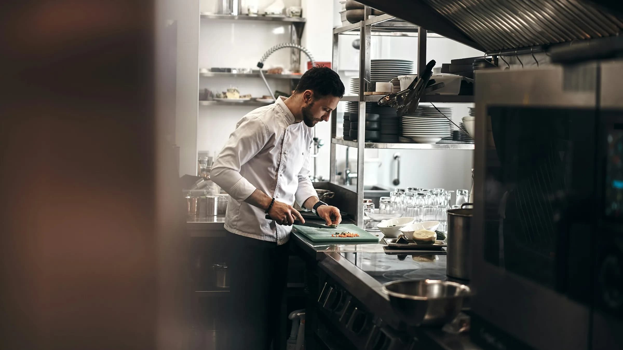 A chef chops ingredients on a board at the steel pass of a restaurant kitchen.