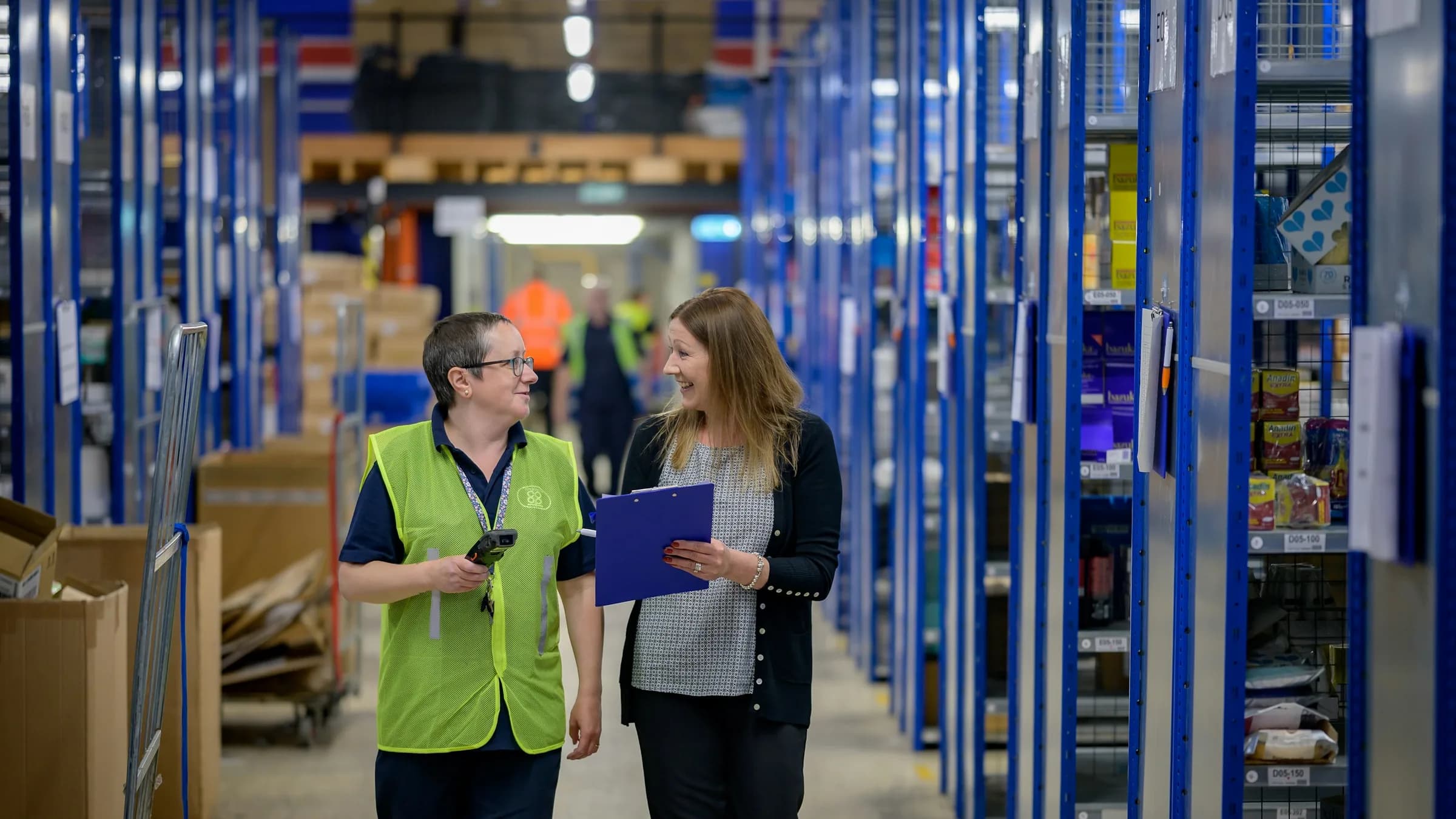 Two staff with a handheld scanner and a clipboard walk down a warehouse aisle of blue shelving.