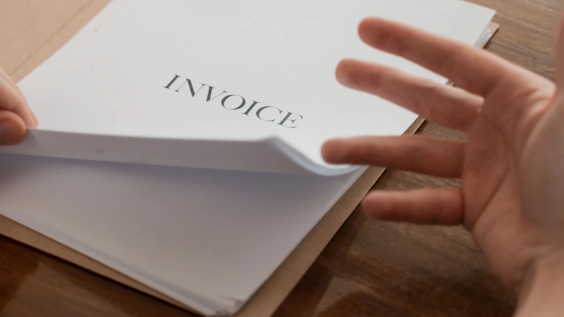 A hand lifts the top sheet of a printed invoice from a folder on a wooden desk.