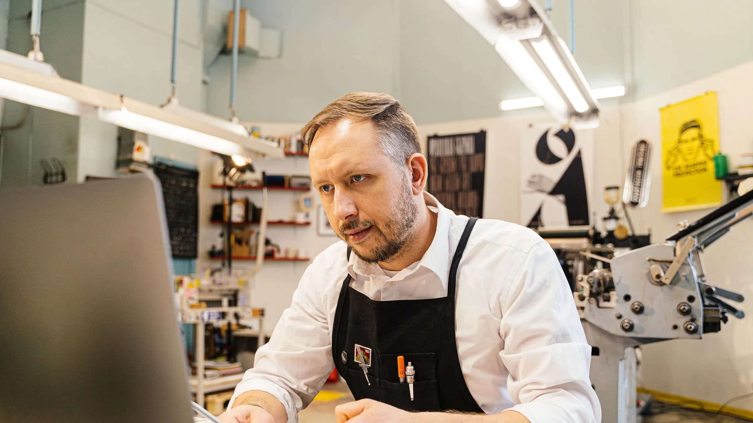 A workshop owner in an apron sits at a computer with a pen and paperwork, machines and shelves behind him.