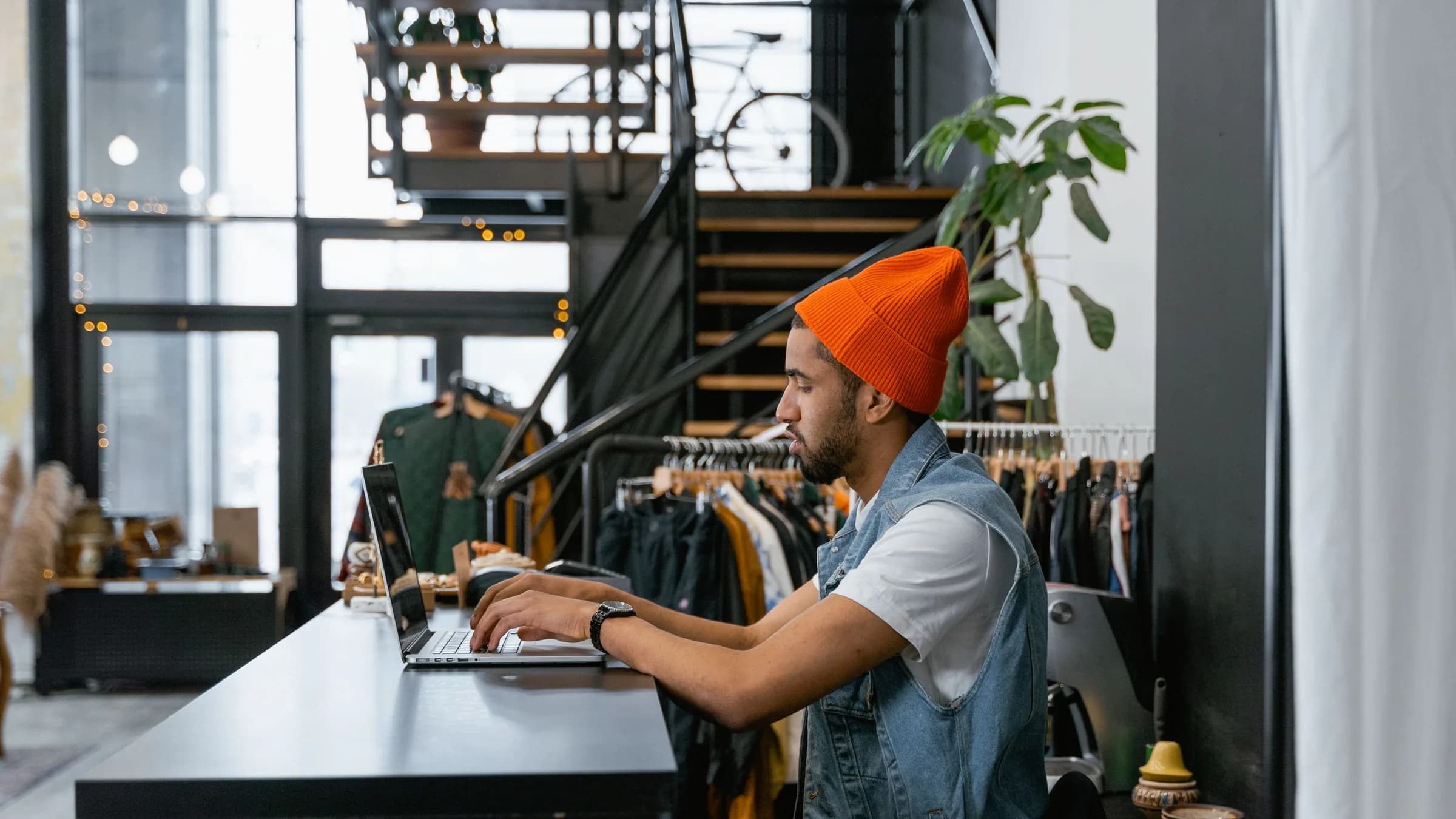 A clothing shop worker types on a laptop at the counter with clothing rails and the shop floor behind him.