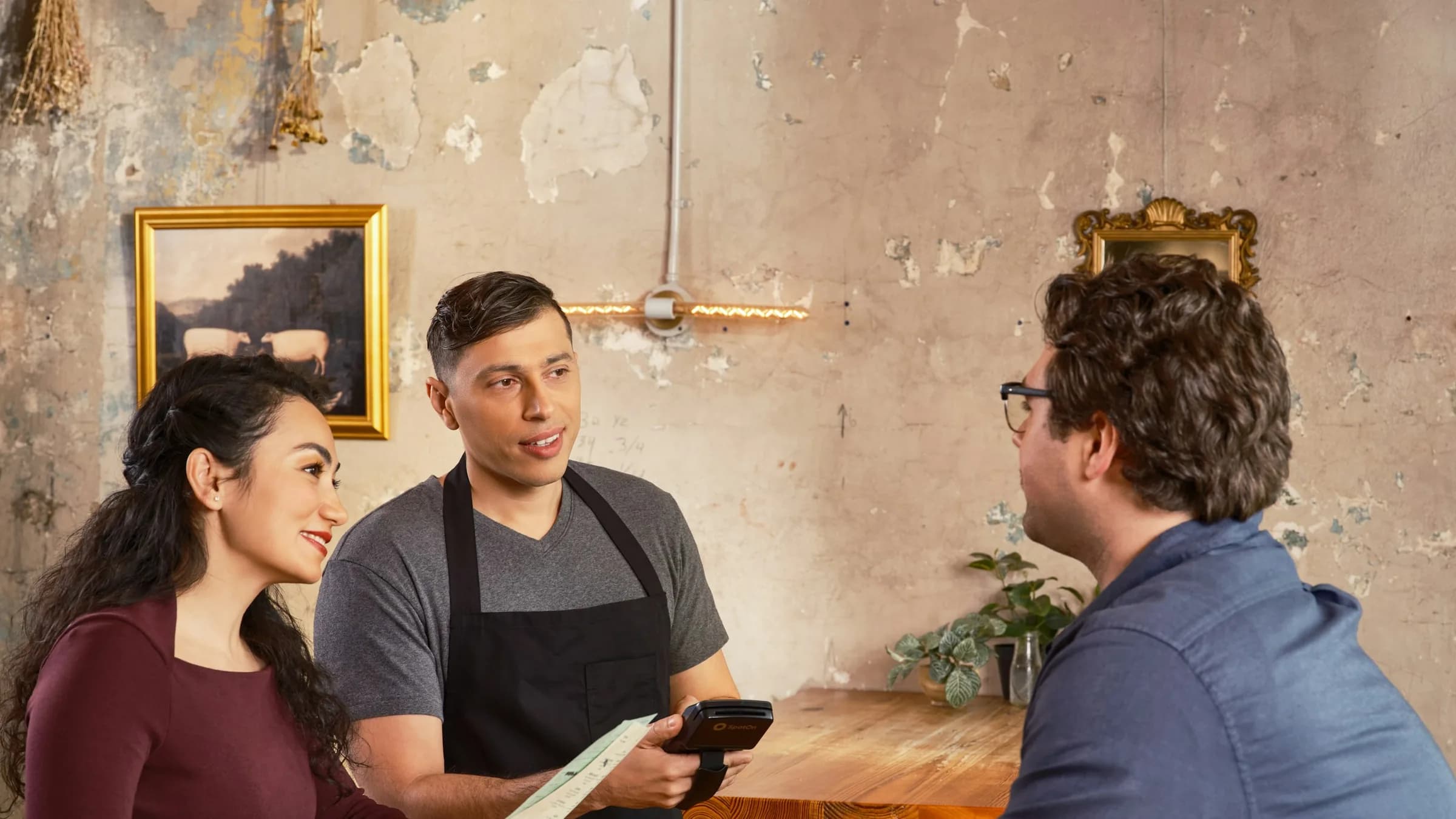 A waiter in an apron takes an order on a handheld device from two diners at a wooden table.