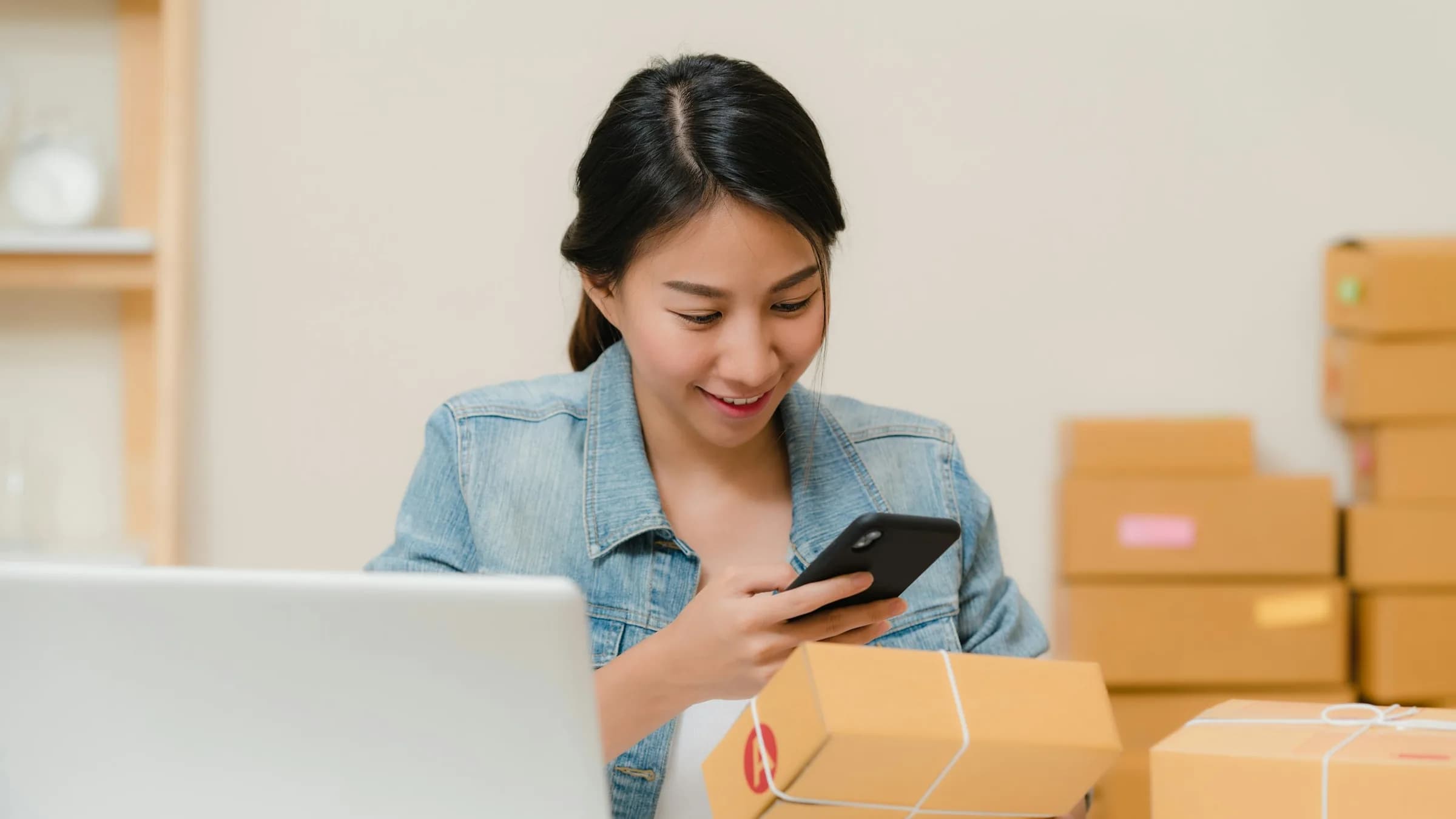 A seller smiles at her phone while holding a parcel, a laptop and a stack of boxes on the table beside her.