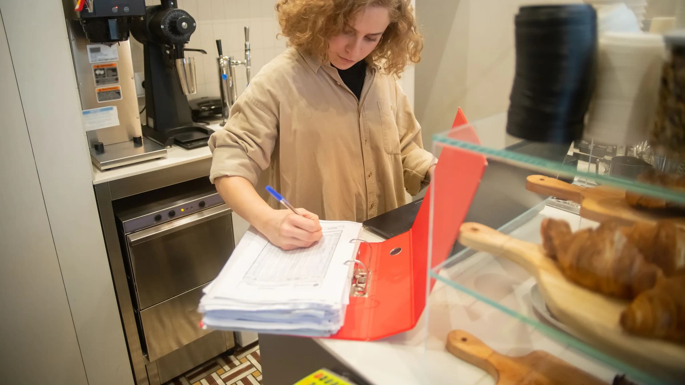 A cafe owner writes in a folder of paperwork at the counter, next to a glass case of pastries and the coffee machine.